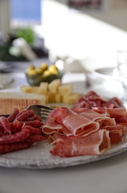 A close-up of a charcuterie board arranged with thin slices of cured meats, cheese cubes, a soft cheese wheel, and some olives in a bowl. The background appears to be a dining setting with bright natural light coming from a window.