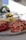 A close-up of a charcuterie board arranged with thin slices of cured meats, cheese cubes, a soft cheese wheel, and some olives in a bowl. The background appears to be a dining setting with bright natural light coming from a window.
