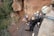 A group of people are geared up for rock climbing or rappelling, standing on a wooden platform attached to a rugged rocky cliff. They are wearing helmets and harnesses, holding onto ropes secured to the rock face. Surrounding them is a dense forest providing a lush green backdrop to the scene.