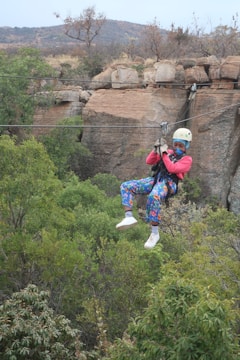 A person wearing a helmet and colorful clothing is zip-lining across a lush, green forest landscape. The background features rocky cliffs and sparse vegetation, with mountains in the distance. The person appears engaged and focused, enjoying an adventurous outdoor activity.
