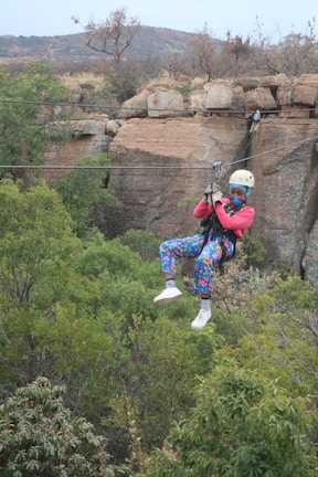 A person wearing a helmet and colorful clothing is zip-lining across a lush, green forest landscape. The background features rocky cliffs and sparse vegetation, with mountains in the distance. The person appears engaged and focused, enjoying an adventurous outdoor activity.