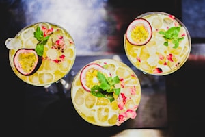 Three cocktail glasses filled with a yellow liquid, ice cubes, and garnished with halved passion fruits, mint leaves, and pink flower petals. The setting is a dark background that contrasts with the vibrant colors of the drinks.