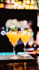 Two elegant cocktail glasses filled with a vibrant orange drink are placed on a bar counter. Each glass is garnished with a small leaf and a decorative element, possibly a slice of fruit or edible flower. The background is intentionally blurred, with bokeh lights creating a warm, inviting atmosphere.