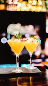 Two elegant cocktail glasses filled with a vibrant orange drink are placed on a bar counter. Each glass is garnished with a small leaf and a decorative element, possibly a slice of fruit or edible flower. The background is intentionally blurred, with bokeh lights creating a warm, inviting atmosphere.