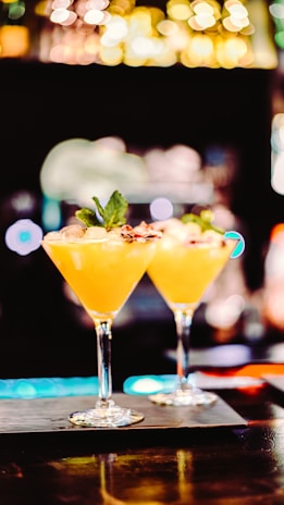 Two elegant cocktail glasses filled with a vibrant orange drink are placed on a bar counter. Each glass is garnished with a small leaf and a decorative element, possibly a slice of fruit or edible flower. The background is intentionally blurred, with bokeh lights creating a warm, inviting atmosphere.