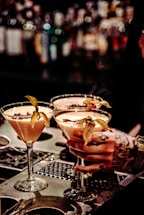 Three elegant cocktails in martini glasses are placed on a bar counter, each garnished with a physalis fruit. A tattooed hand is holding one of the glasses, suggesting an atmosphere of sophistication and style. The background is a blurred display of numerous liquor bottles, creating a warm and inviting ambiance.