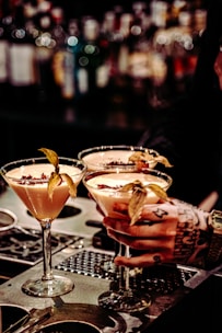 Three elegant cocktails in martini glasses are placed on a bar counter, each garnished with a physalis fruit. A tattooed hand is holding one of the glasses, suggesting an atmosphere of sophistication and style. The background is a blurred display of numerous liquor bottles, creating a warm and inviting ambiance.