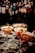 Three elegant cocktails in martini glasses are placed on a bar counter, each garnished with a physalis fruit. A tattooed hand is holding one of the glasses, suggesting an atmosphere of sophistication and style. The background is a blurred display of numerous liquor bottles, creating a warm and inviting ambiance.