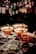 Three elegant cocktails in martini glasses are placed on a bar counter, each garnished with a physalis fruit. A tattooed hand is holding one of the glasses, suggesting an atmosphere of sophistication and style. The background is a blurred display of numerous liquor bottles, creating a warm and inviting ambiance.