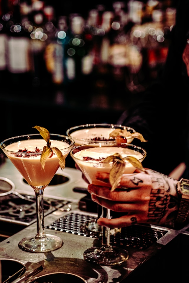 Three elegant cocktails in martini glasses are placed on a bar counter, each garnished with a physalis fruit. A tattooed hand is holding one of the glasses, suggesting an atmosphere of sophistication and style. The background is a blurred display of numerous liquor bottles, creating a warm and inviting ambiance.
