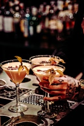 Three elegant cocktails in martini glasses are placed on a bar counter, each garnished with a physalis fruit. A tattooed hand is holding one of the glasses, suggesting an atmosphere of sophistication and style. The background is a blurred display of numerous liquor bottles, creating a warm and inviting ambiance.