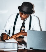 A person wearing a hat and glasses is seated at a desk, writing with a pen on a piece of paper. They are wearing a white shirt, black suspenders, and a patterned tie. The desk is cluttered with a laptop, a magazine or newspaper, and a whiteboard or notepad.