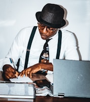 A person wearing a hat and glasses is seated at a desk, writing with a pen on a piece of paper. They are wearing a white shirt, black suspenders, and a patterned tie. The desk is cluttered with a laptop, a magazine or newspaper, and a whiteboard or notepad.