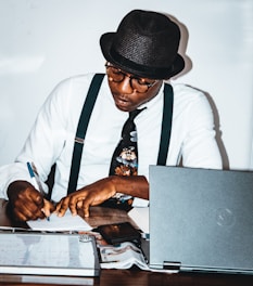 A person wearing a hat and glasses is seated at a desk, writing with a pen on a piece of paper. They are wearing a white shirt, black suspenders, and a patterned tie. The desk is cluttered with a laptop, a magazine or newspaper, and a whiteboard or notepad.