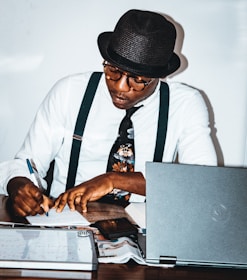 A person wearing a hat and glasses is seated at a desk, writing with a pen on a piece of paper. They are wearing a white shirt, black suspenders, and a patterned tie. The desk is cluttered with a laptop, a magazine or newspaper, and a whiteboard or notepad.