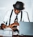 A person wearing a hat and glasses is seated at a desk, writing with a pen on a piece of paper. They are wearing a white shirt, black suspenders, and a patterned tie. The desk is cluttered with a laptop, a magazine or newspaper, and a whiteboard or notepad.