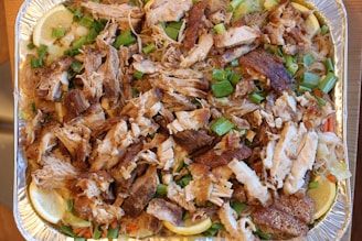 A tray filled with cooked shredded meat, likely chicken or pork, mixed with vermicelli noodles. The dish is garnished with chopped green onions, slices of lemon, and includes some vegetables such as carrots and cabbage. The meat appears to have a crispy texture.