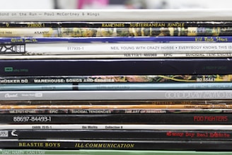 A stack of vinyl record spines featuring various albums from different musical artists. The text on each spine varies, displaying album and artist names along with other numeric or label information. The records are stacked in a slightly uneven manner, showcasing a collection with artistic and nostalgic appeal.