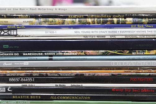 A stack of vinyl record spines featuring various albums from different musical artists. The text on each spine varies, displaying album and artist names along with other numeric or label information. The records are stacked in a slightly uneven manner, showcasing a collection with artistic and nostalgic appeal.