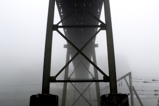 A structural bridge framework towers into the foggy air, with dense mist obscuring the background. The construction features steel beams intersecting in a geometric pattern. Below, electrical wires stretch horizontally, adding complexity to the scene.