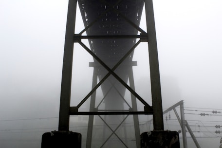A structural bridge framework towers into the foggy air, with dense mist obscuring the background. The construction features steel beams intersecting in a geometric pattern. Below, electrical wires stretch horizontally, adding complexity to the scene.