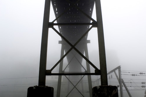 A structural bridge framework towers into the foggy air, with dense mist obscuring the background. The construction features steel beams intersecting in a geometric pattern. Below, electrical wires stretch horizontally, adding complexity to the scene.