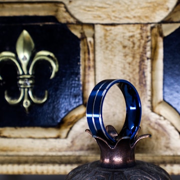A shiny metallic blue ring with a dual stripe design is elegantly balanced on the pointed end of a decorative object. In the background, a wooden plaque with a gold fleur-de-lis symbol is visible, providing a vintage and sophisticated ambiance.