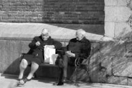 Two elderly people sit on a bench against a brick and stone wall. One person is going through a shopping bag while the other sits calmly with hands resting on their lap. The scene is in black and white.