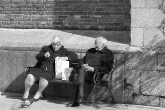 Two elderly people sit on a bench against a brick and stone wall. One person is going through a shopping bag while the other sits calmly with hands resting on their lap. The scene is in black and white.