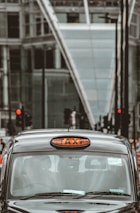 A classic black taxi with a prominent 'TAXI' sign on top is captured in an urban setting. The background consists of modern glass buildings and traffic lights, likely in a bustling city environment.