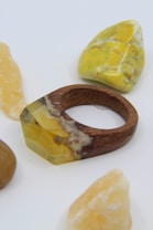 A wooden ring featuring a resin component with yellow and brown inclusions lies amid several polished stones, predominantly yellow and brown in color.