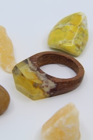 A wooden ring featuring a resin component with yellow and brown inclusions lies amid several polished stones, predominantly yellow and brown in color.