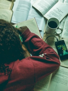 A person wearing a maroon hoodie is writing or drawing in a notebook surrounded by several open books. There is also a mug filled with a dark beverage, a pair of glasses, and a smartphone displaying an image or video.