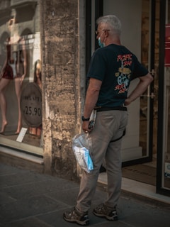 An older man with grey hair is standing in front of a storefront window displaying mannequins dressed in lingerie. He is wearing a black T-shirt with a graphic on the back, beige pants, and a wristwatch. He's holding a plastic bag containing an unopened mask in his left hand and has a face mask pulled down under his chin.