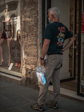 An older man with grey hair is standing in front of a storefront window displaying mannequins dressed in lingerie. He is wearing a black T-shirt with a graphic on the back, beige pants, and a wristwatch. He's holding a plastic bag containing an unopened mask in his left hand and has a face mask pulled down under his chin.