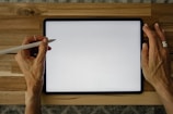 A person holds a digital stylus near a tablet with a blank white screen, placed on a wooden surface. Both hands are visible, poised over the tablet.