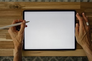 A person holds a digital stylus near a tablet with a blank white screen, placed on a wooden surface. Both hands are visible, poised over the tablet.