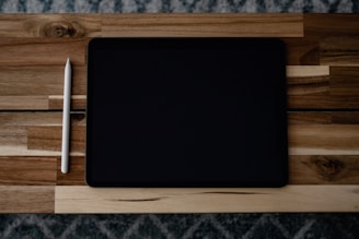 A tablet and a stylus placed on a wooden table with a textured pattern. The tablet has a black screen and is positioned horizontally, while the white stylus is placed vertically to its left.