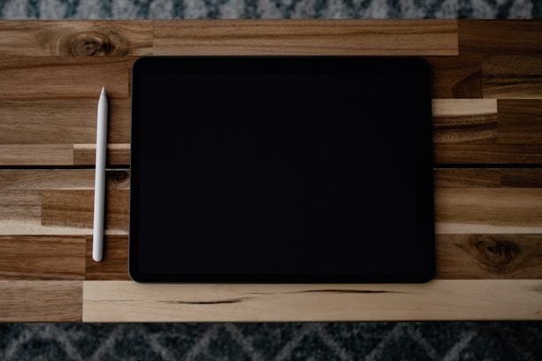 A tablet and a stylus placed on a wooden table with a textured pattern. The tablet has a black screen and is positioned horizontally, while the white stylus is placed vertically to its left.