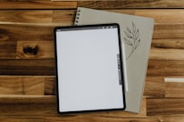 A digital tablet with a blank screen is placed on top of a closed notebook adorned with a leaf illustration. A stylus rests between the tablet and the notebook, all situated on a wooden surface.