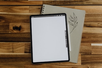 A digital tablet with a blank screen is placed on top of a closed notebook adorned with a leaf illustration. A stylus rests between the tablet and the notebook, all situated on a wooden surface.
