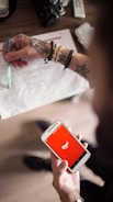 A person with tattoos on their arms is holding a smartphone displaying the iFood app against a red background. In the other hand, they are holding a pen over a printed document. The scene suggests multitasking or working while using a food delivery app. Several accessories like a bracelet and ring are visible, and a camera and remote control are placed on the table.