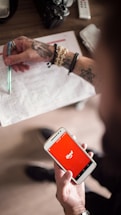 A person with tattoos on their arms is holding a smartphone displaying the iFood app against a red background. In the other hand, they are holding a pen over a printed document. The scene suggests multitasking or working while using a food delivery app. Several accessories like a bracelet and ring are visible, and a camera and remote control are placed on the table.