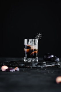 A tall glass filled with a dark beverage, garnished with a sprig of rosemary, stands on a dark surface. Around the glass, sliced purple fruit and additional sprigs are artistically scattered. The background is a deep black, emphasizing the rich colors of the drink and garnish.