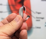 A close-up of a hand holding a small hearing aid. The background is blurred, possibly showing some educational or instructional material.