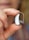 A close-up of a hand holding a small hearing aid between the thumb and index finger. The hearing aid is sleek and metallic, with a clear tube extending from it. The background is softly blurred, emphasizing the focus on the hearing aid.