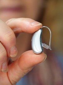 A close-up of a hand holding a small hearing aid between the thumb and index finger. The hearing aid is sleek and metallic, with a clear tube extending from it. The background is softly blurred, emphasizing the focus on the hearing aid.