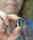 A close-up of a hand holding a small hearing aid device between the thumb and forefinger. The hearing aid is sleek and modern, predominantly black and silver with a clear plastic tube.