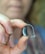 A close-up of a hand holding a small hearing aid device between the thumb and forefinger. The hearing aid is sleek and modern, predominantly black and silver with a clear plastic tube.