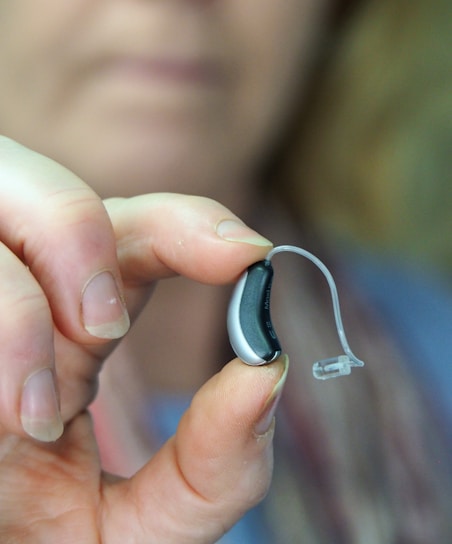 A close-up of a hand holding a small hearing aid device between the thumb and forefinger. The hearing aid is sleek and modern, predominantly black and silver with a clear plastic tube.